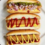 three vegan hot dogs in buns on a plater with toppings