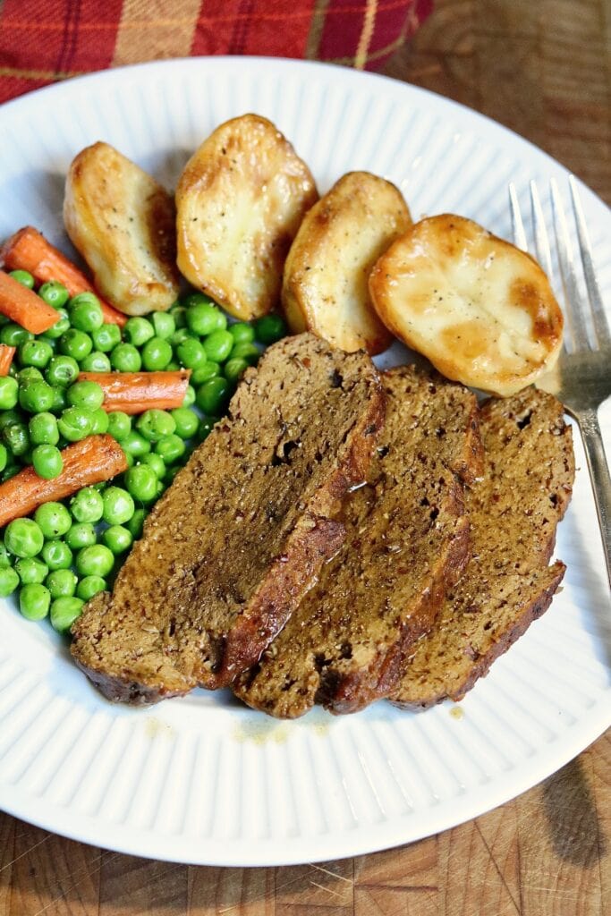 vegan roast beef sliced on a plate with roasted potatoes and peas