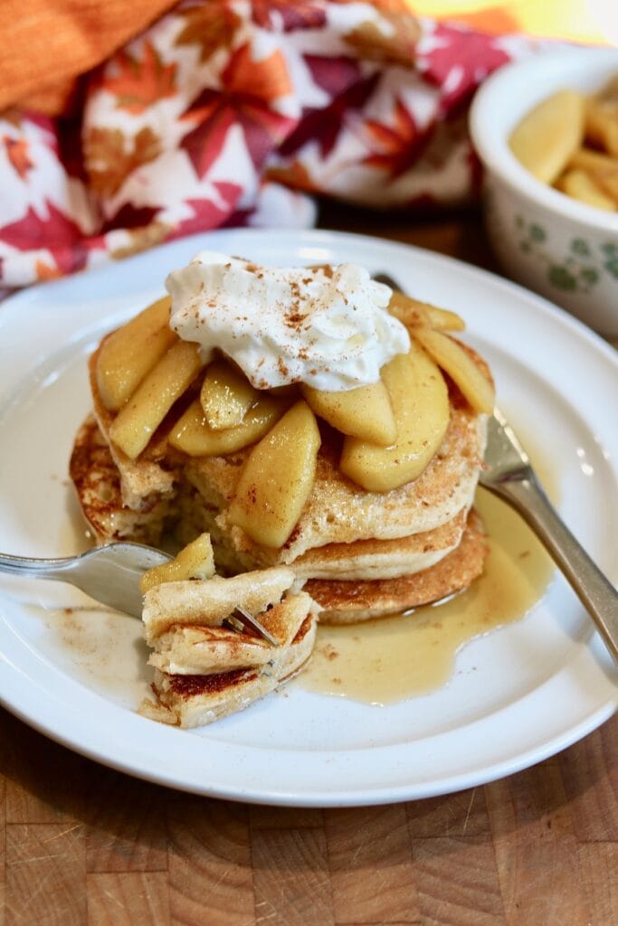 vegan apple cinnamon pancakes stacked on a plate with apple topping being sliced with fork and knife