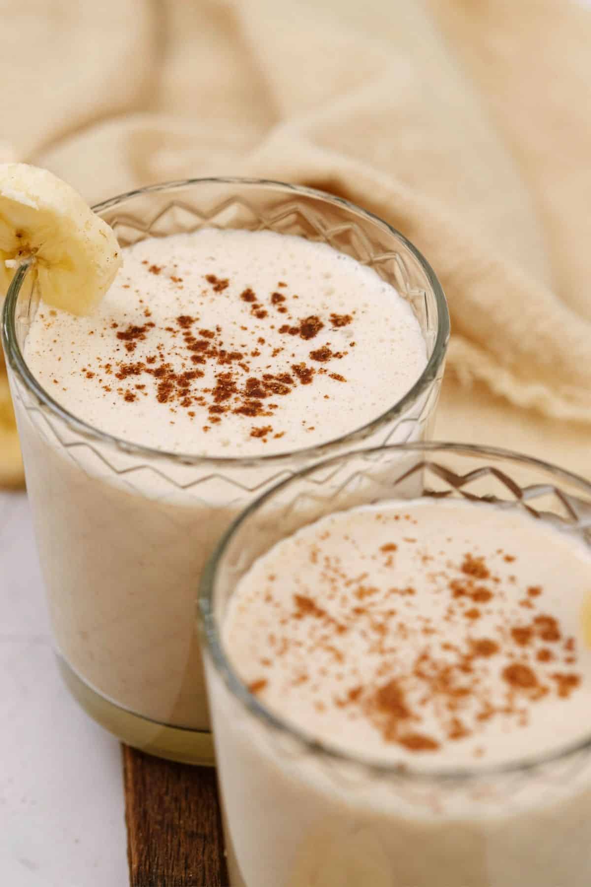 two short glasses of oat smoothie with peanut butter topped with sprinkle of cinnamon and banana slice
