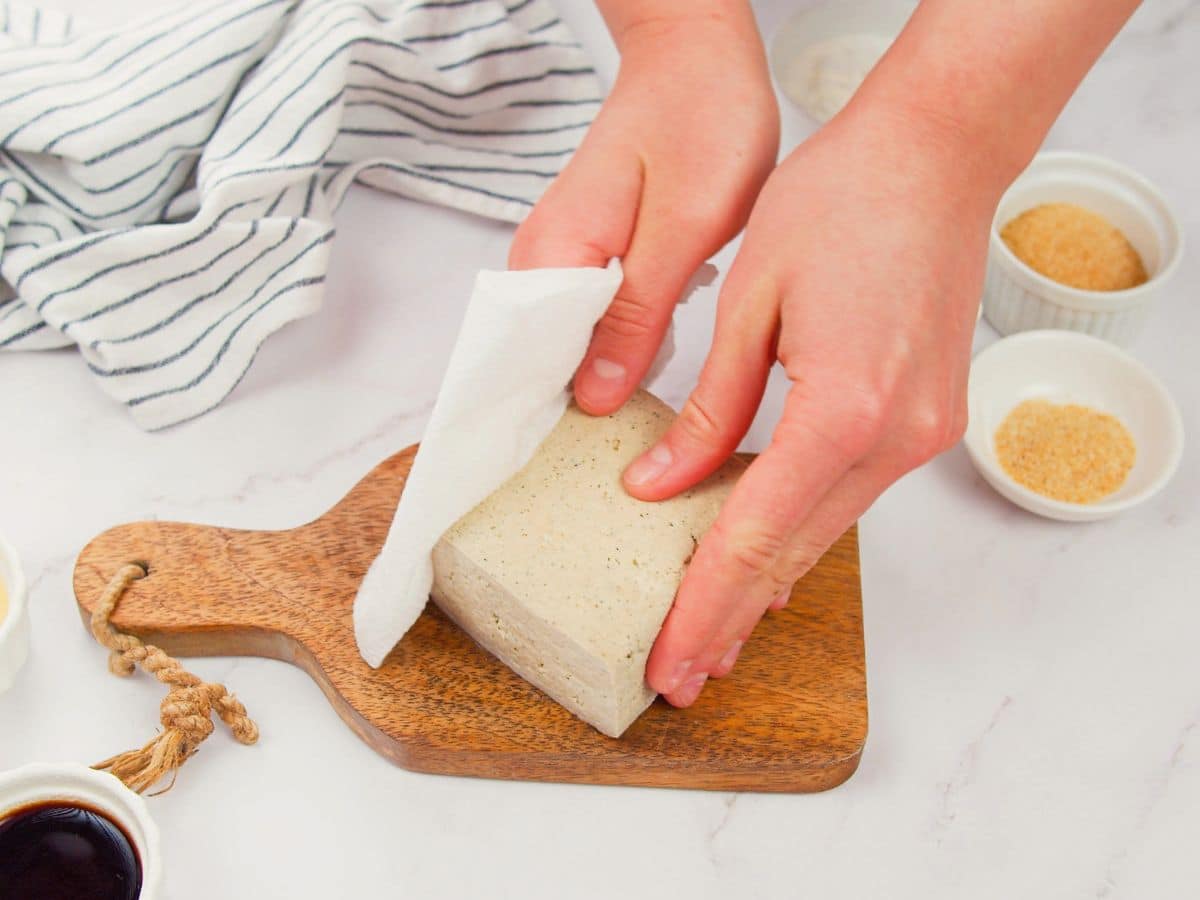 paper towel patting side of tofu block on cutting board on white table