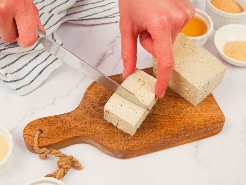 hand holding block of tofu while cutting on small board