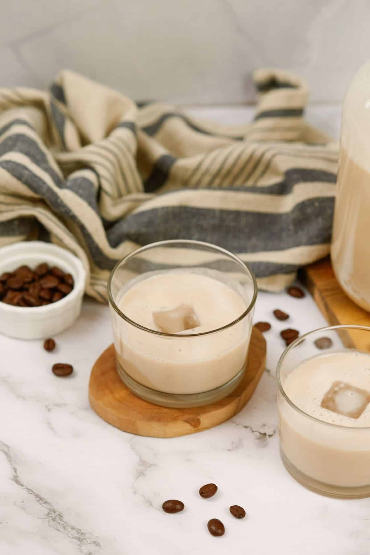 two glasses of vegan Bailey's Irish cream on wood coasters on table by striped towel