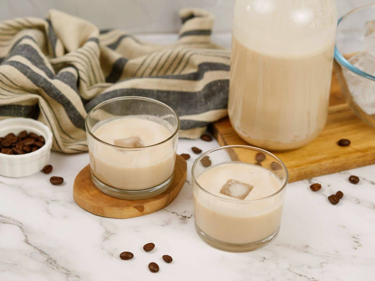 striped towel behind wood board holding jar of cream behind two short cocktail glasses of vegan Bailey's