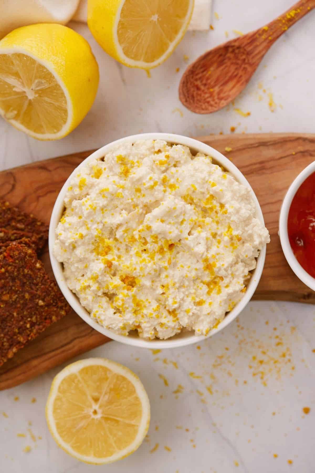 round white bowl of vegan tofu cottage cheese nexxt to bowl of jam on board next to sliced lemons