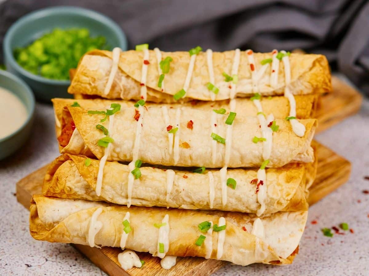 gray napkin and bowl of fresh onions in background behind stack of baked taquitos