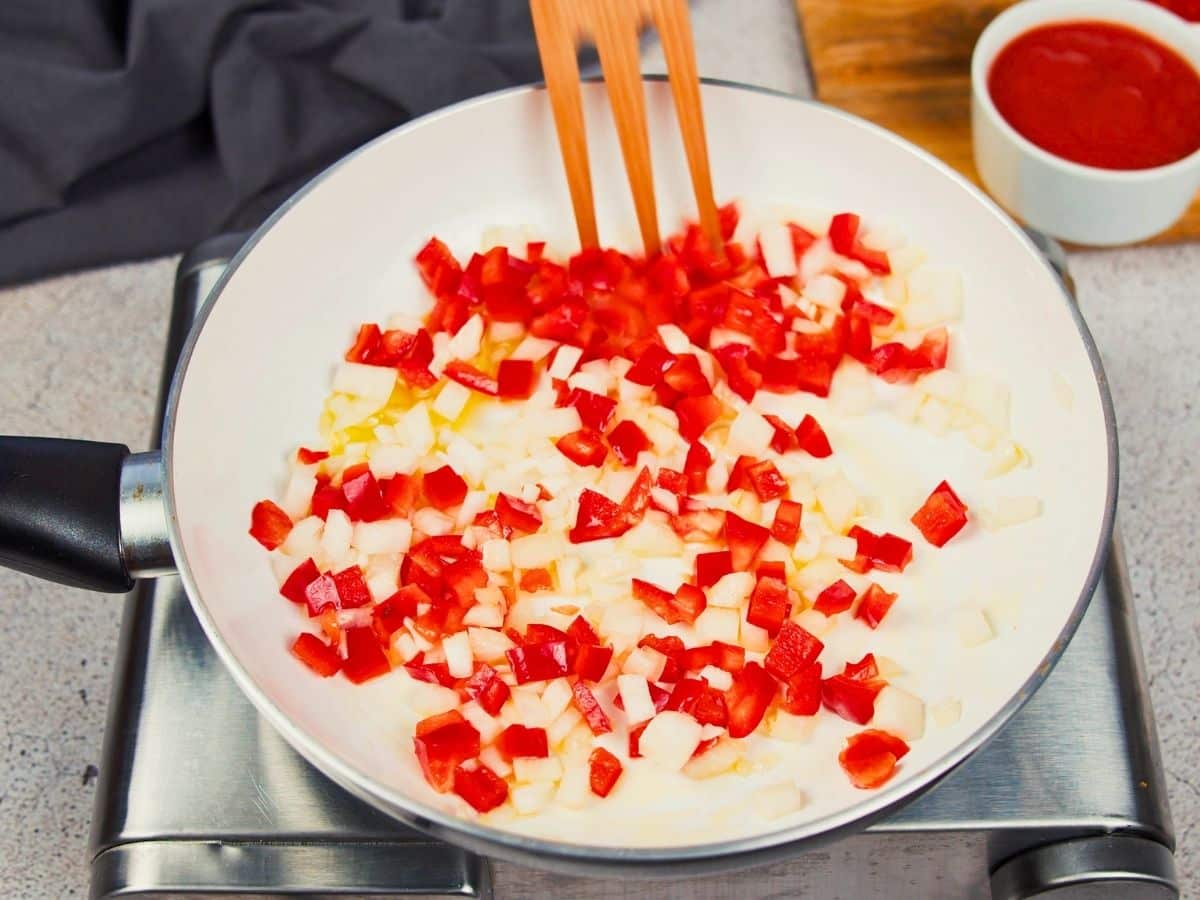 wooden spoon in white skillet with onions and red bell peppers