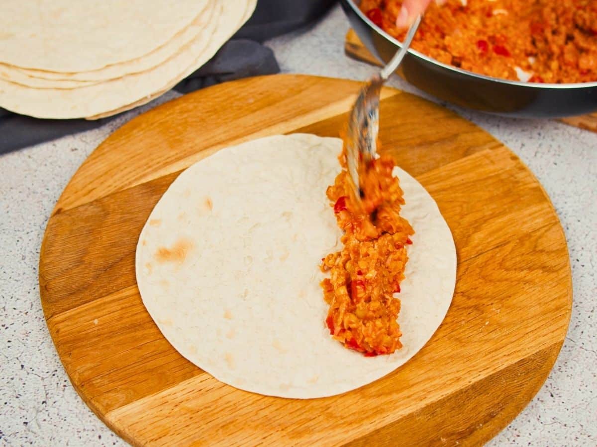 hand putting spoon of buffalo chickpea mixture onto tortilla on cutting board
