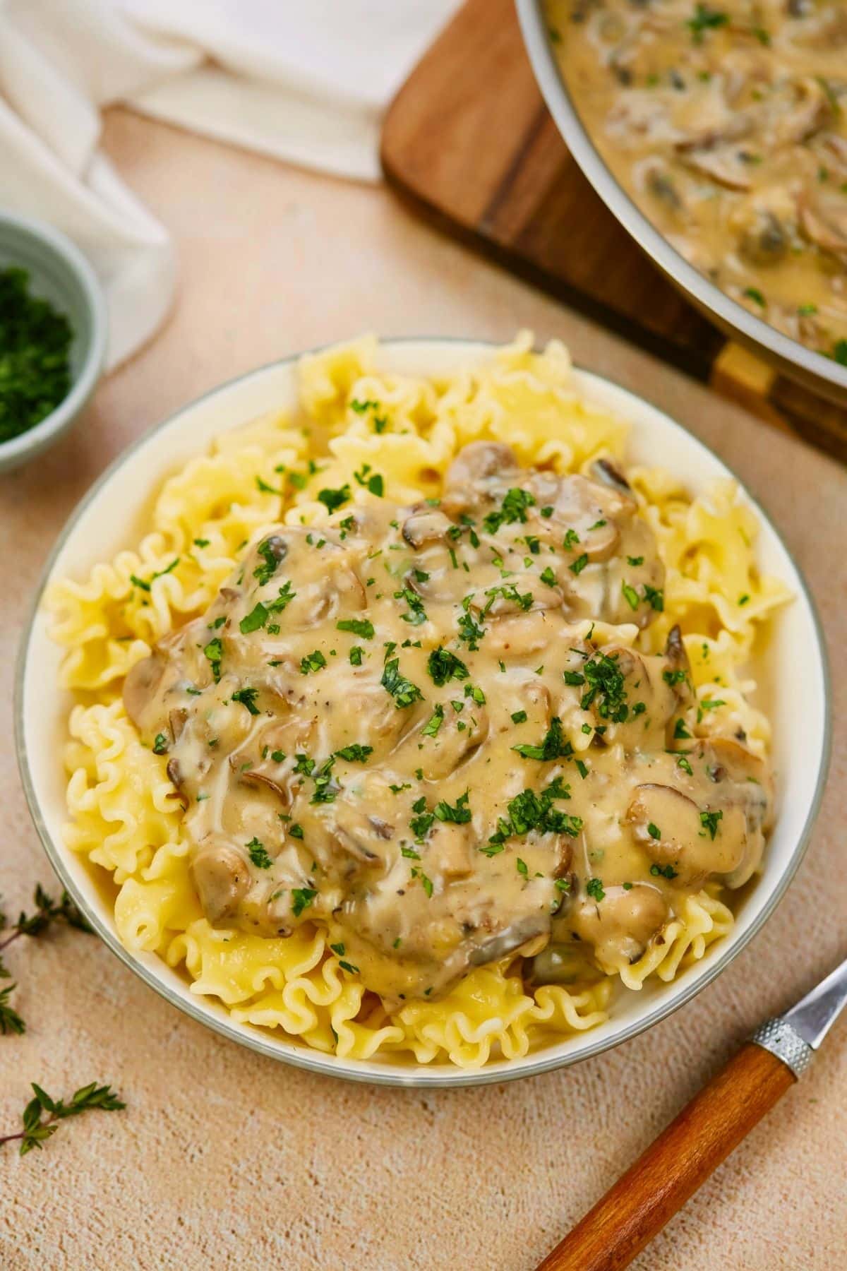 round bowl of vegan mushroom stroganoff on table by wooden bottom spoon and skillet of mushroom sauce