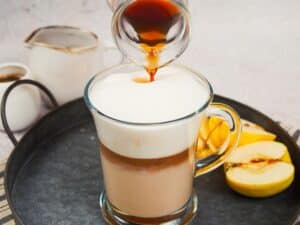 syrup being poured over foam on top of apple crisp macchiato in glass mug on gray platter