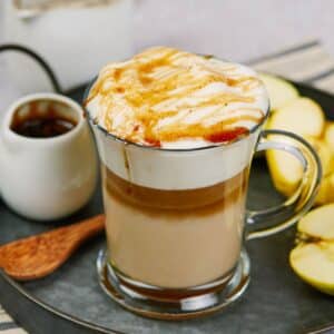 glass mug of layered coffee drink topped by foam and drizzle of syrup sitting on gray tray