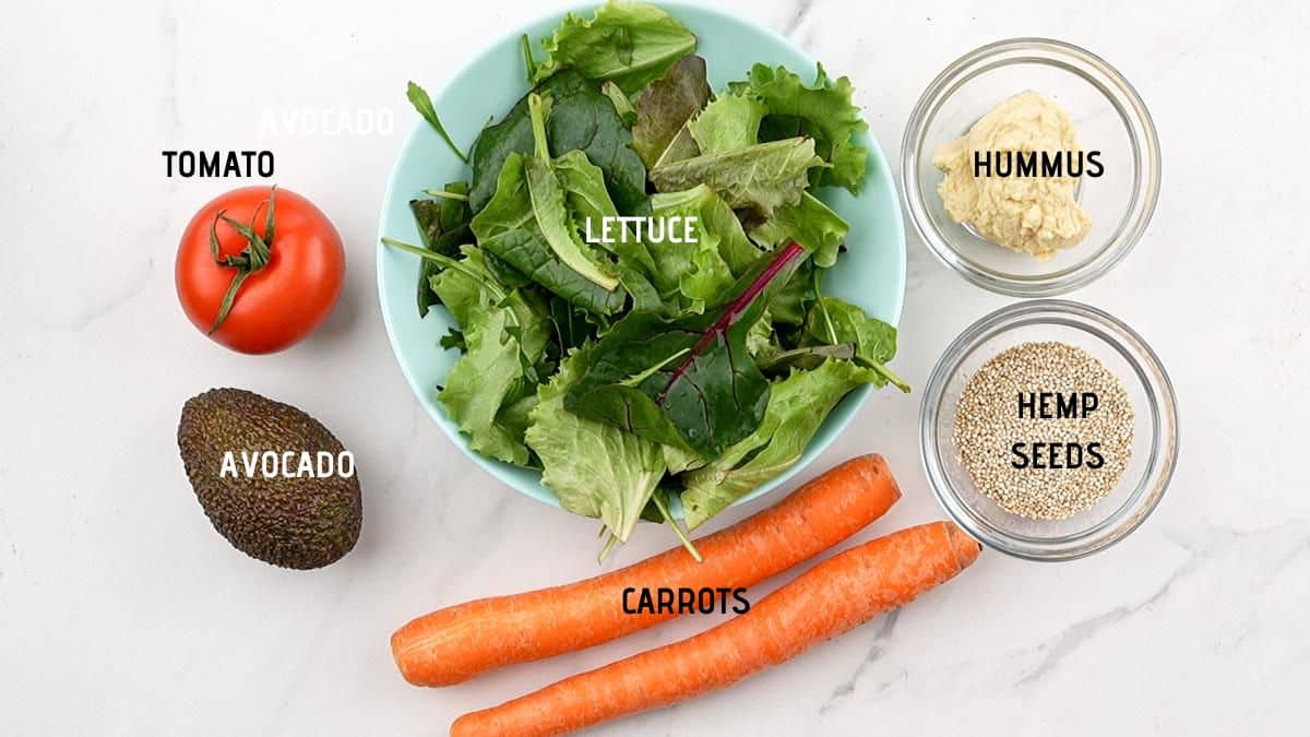 blue bowl of lettuce on table with tomato, avocado, carrots, and bowls of ingredients on white table