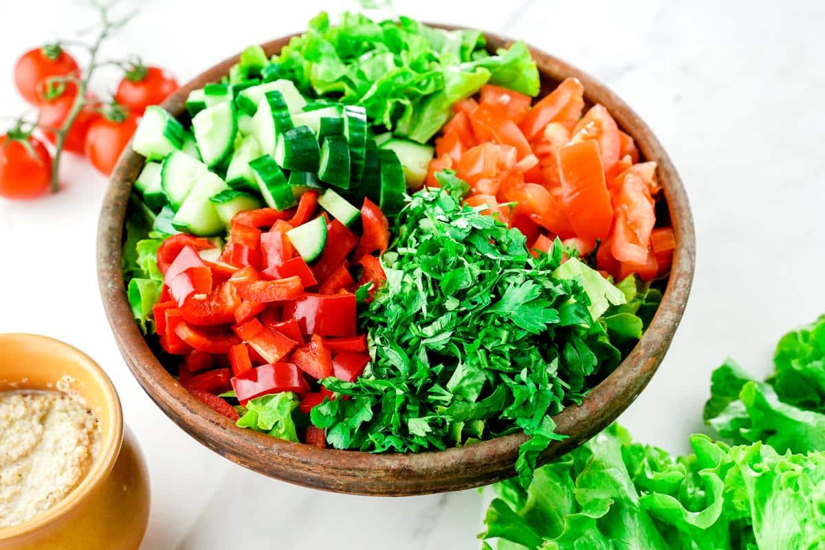 large wooden bowl of lettuce, tomato, peppers, cucumbers, and herbs on whtie table