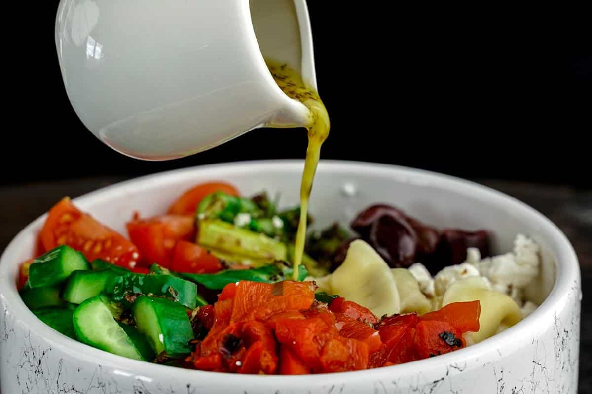 dressing being poured over greek tortellini pasta salad in large white bowl