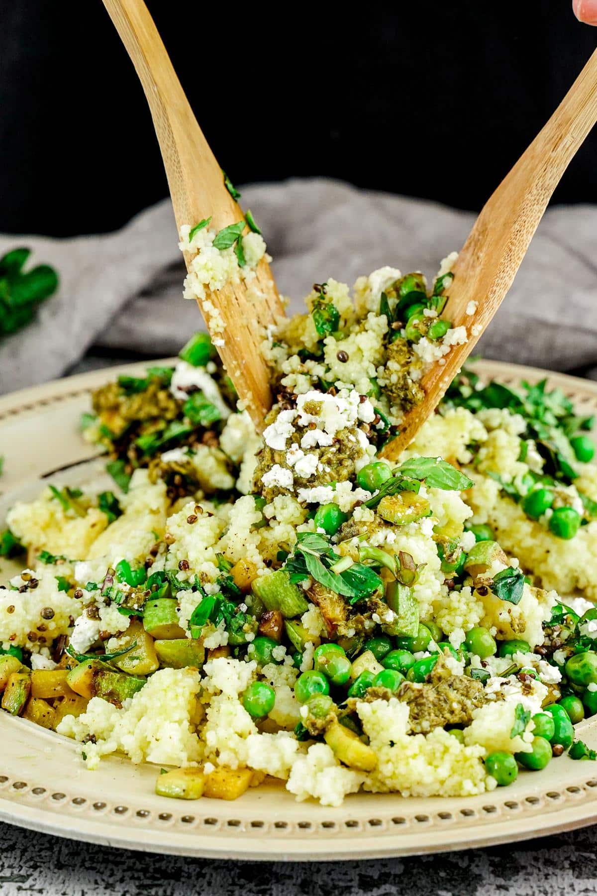 wooden spoons tossing pesto couscous salad above cream plate with gold rim