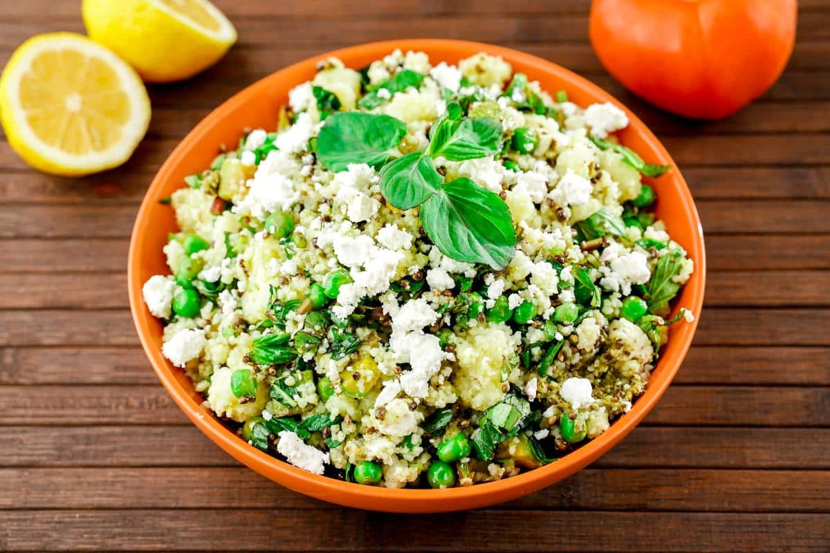 orange bowl of pesto couscous salad on wood table by lemon halves and tomato