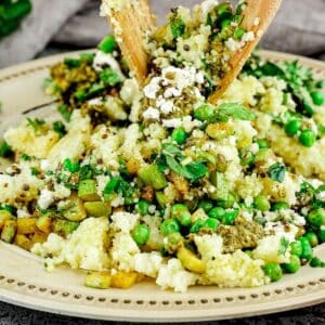 wooden spoons tossing pesto couscous salad above cream plate with gold rim
