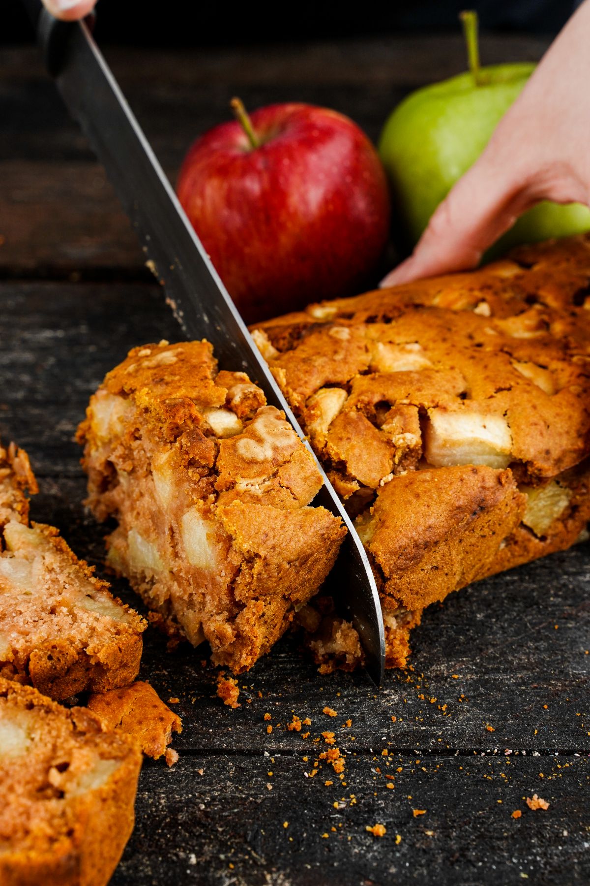 apple bread sitting on black counter by apples being sliced with large knife