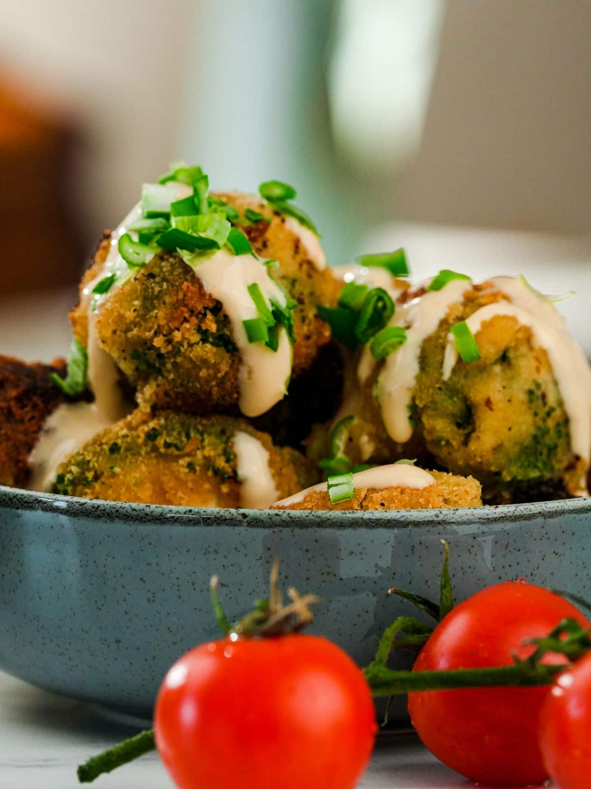 blue bowl of bang bang broccoli topped with sauce and chives on table by tomatoes