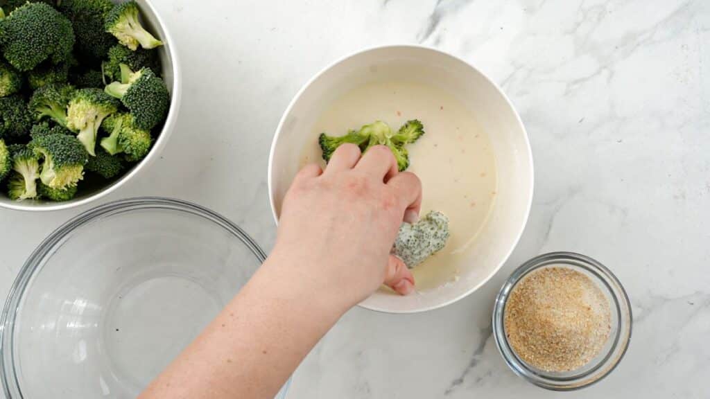 hand dipping broccoli floret into white bowl of batter