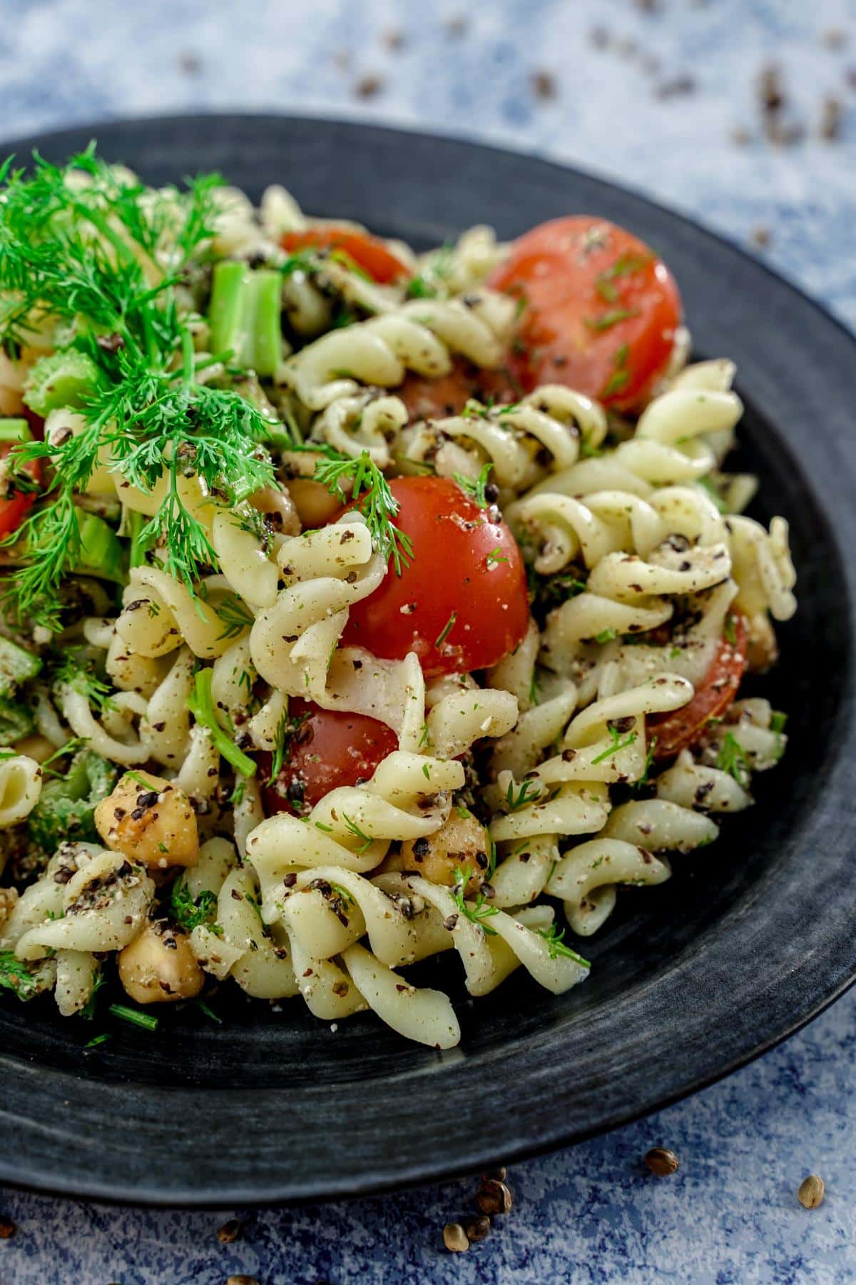 black plate on blue table topped with pasta salad with tomato and dill