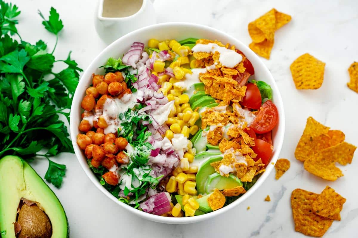 round white bowl of bbq chickpea chopped salad on table next to chips, avocado, and herbs