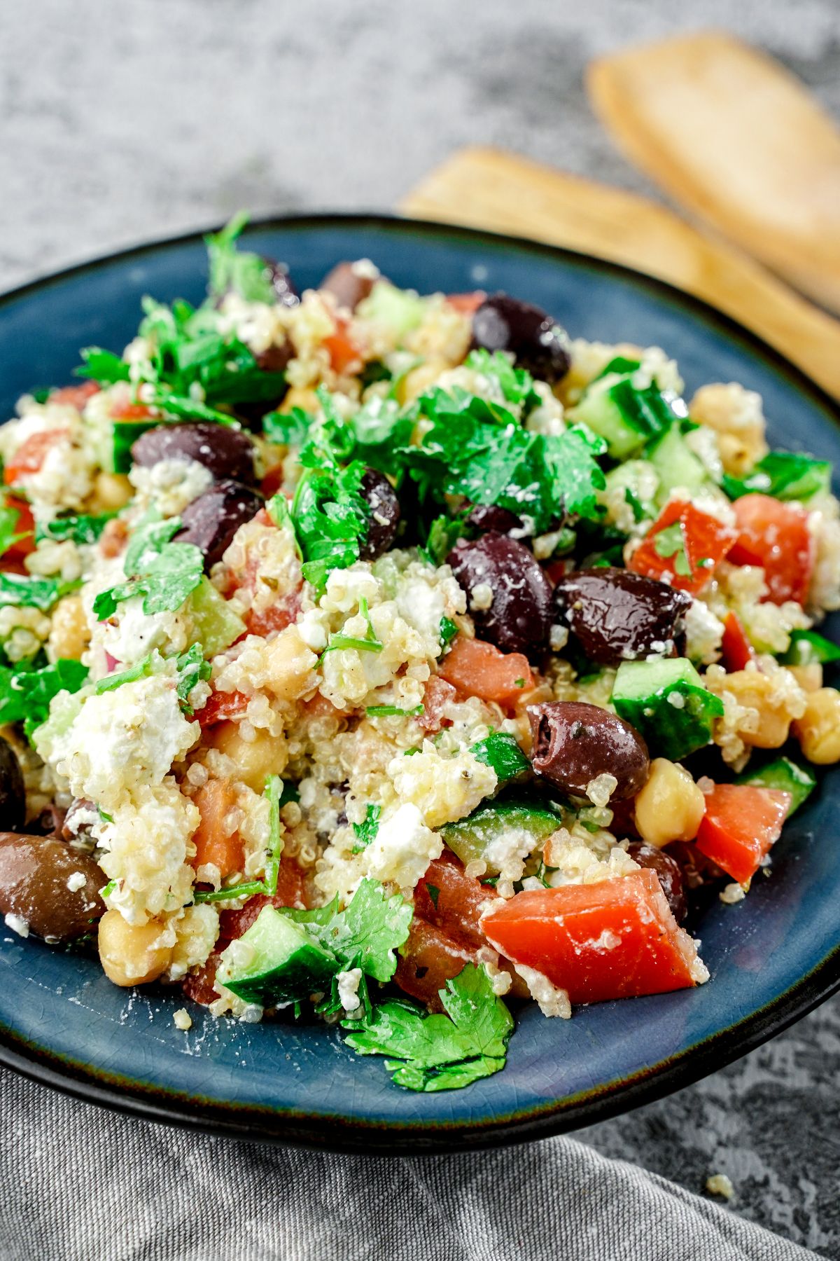 dark blue plate of Greek quinoa salad on gray tablecloth with wooden spoon in background