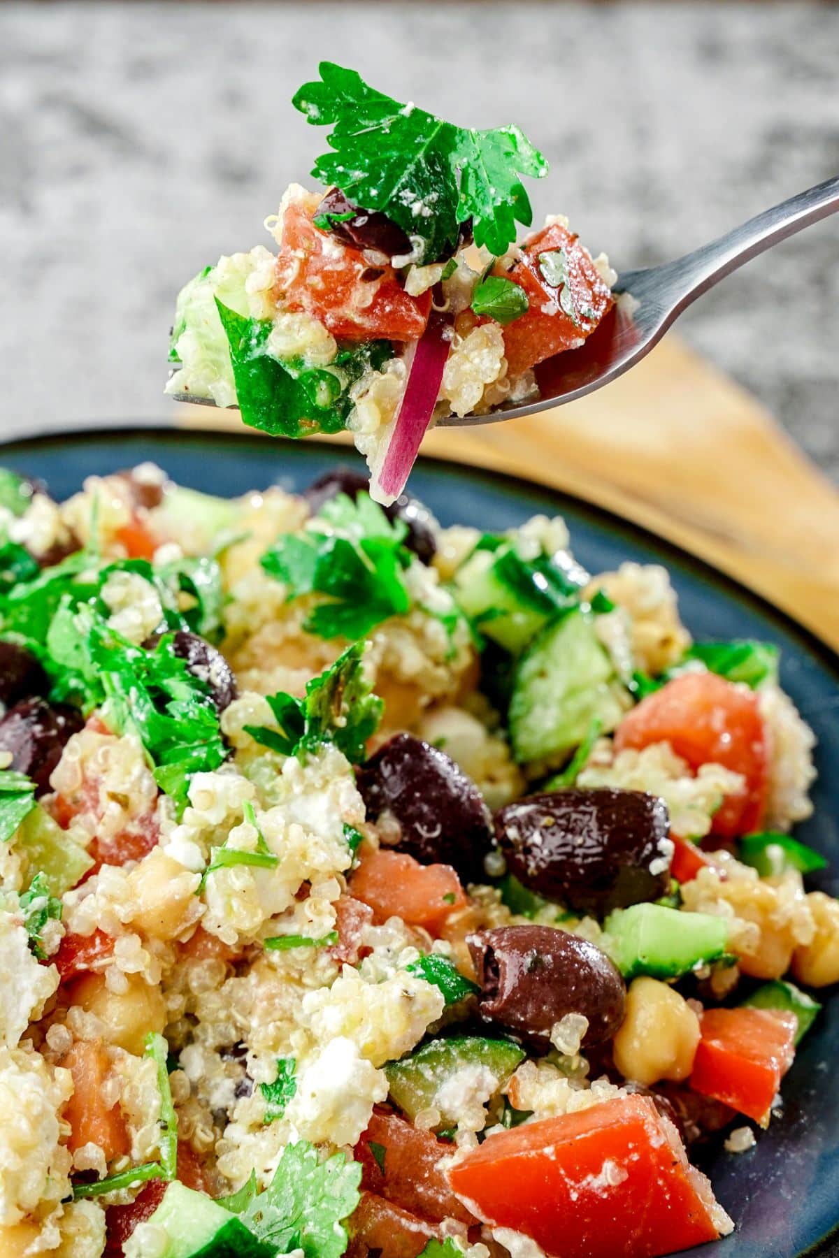 blue plate of Greek quinoa salad with fork held above plate and wooden spoon in background