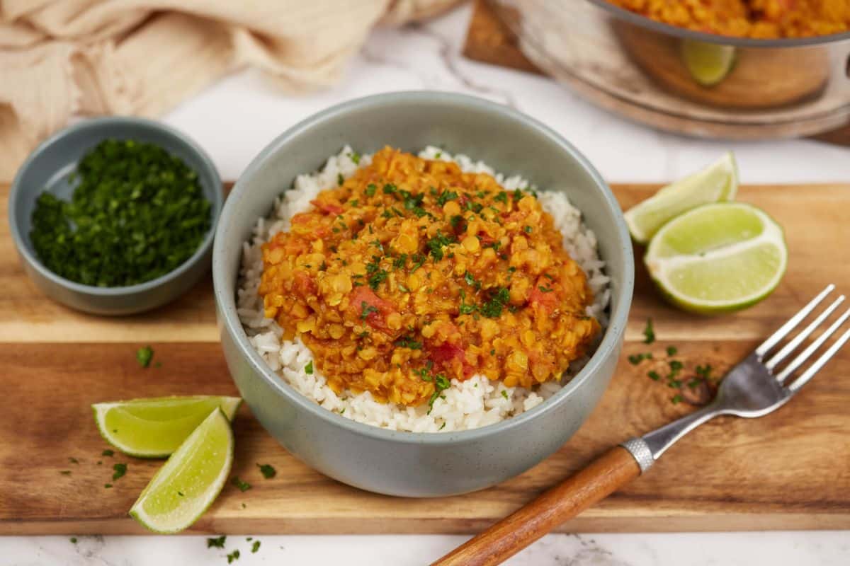 wooden cutting board holding gray bowl of rice and red lentil dahl with lime wedges on the side