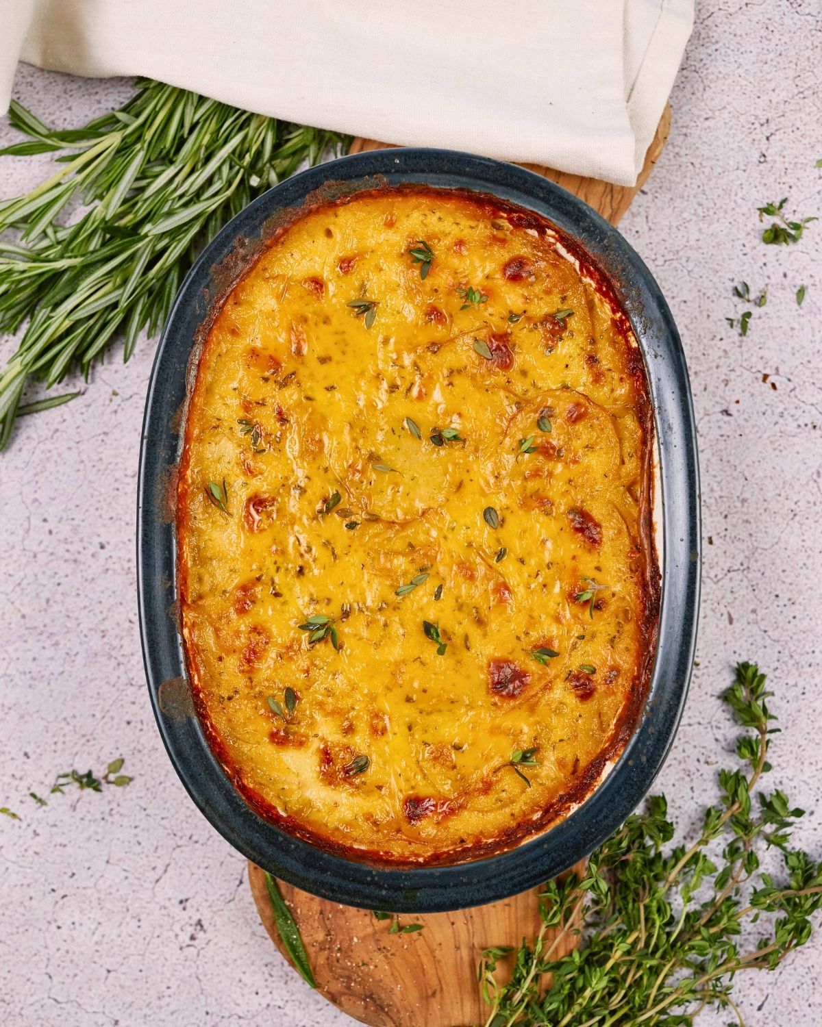 blue and white oval baking dish of vegan scalloped potatoes on table by fresh herbs