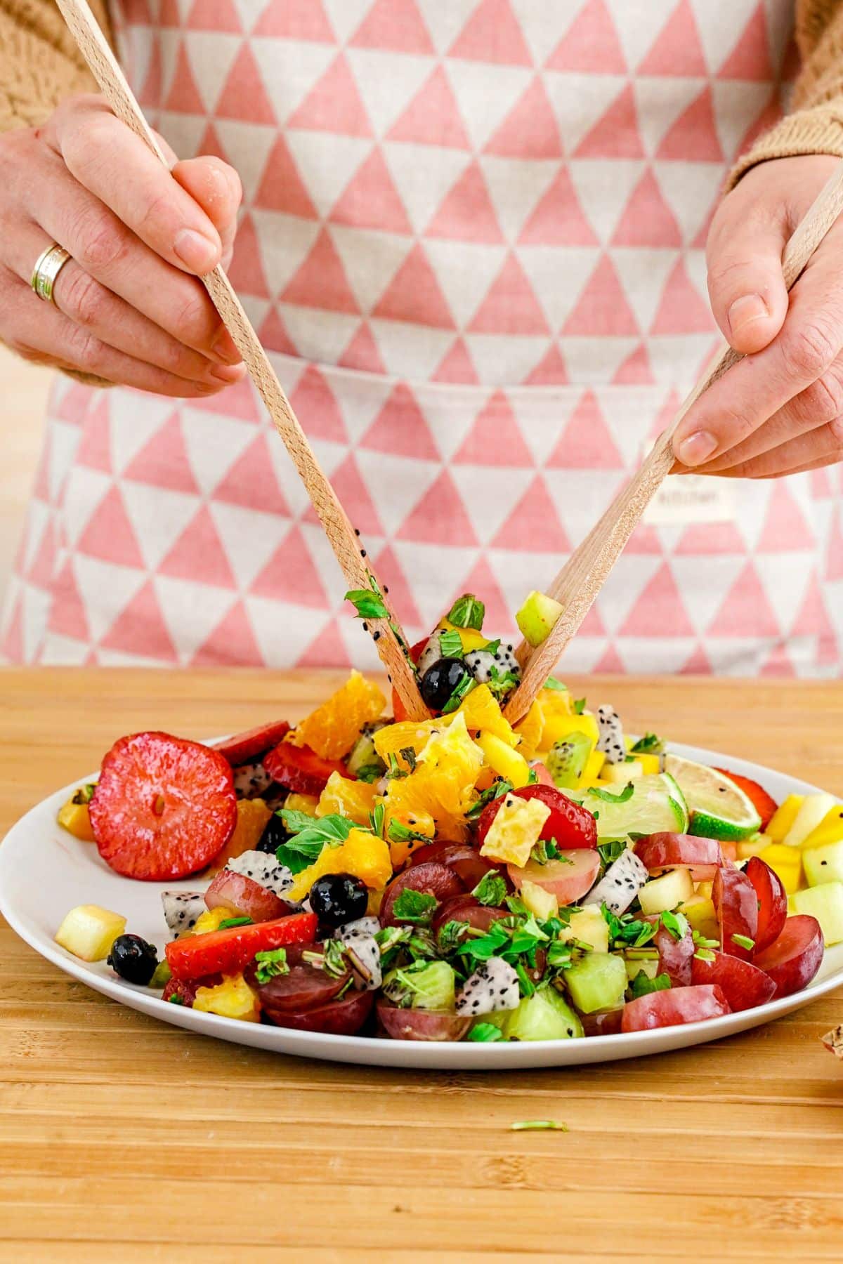 woman using wooden spoons to toss summer fruit salad on plate