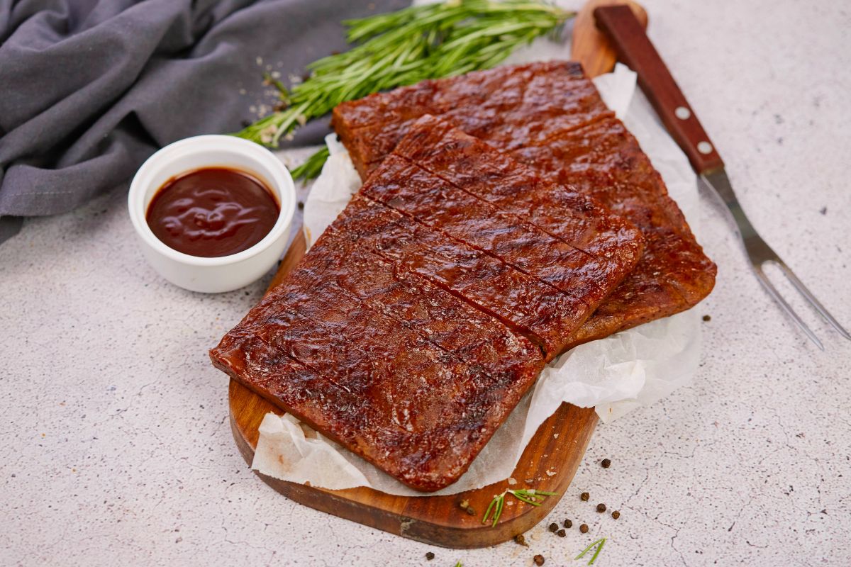 sliced vegan bbq ribs on platter by bowl of sauce and fresh herbs